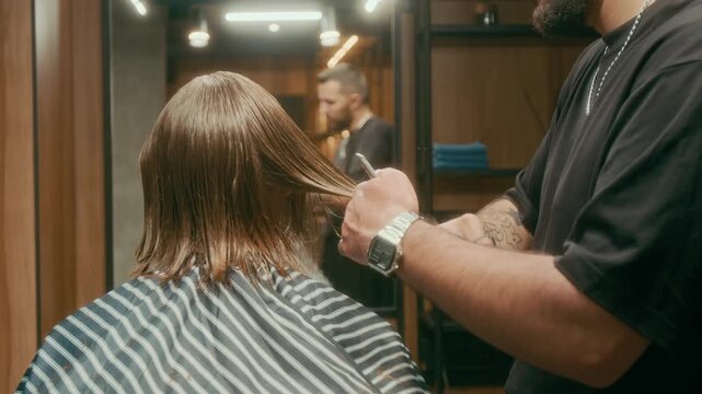 Medium shot of bearded skillful young barber tapering clients red hair to achieve desired shape and volume, while working on haircut in barbershop with wooden wall panels and mirrors