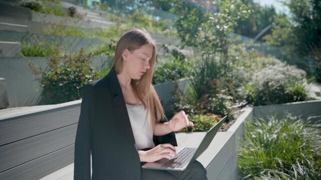 Busy blonde woman freelancer working on laptop placed on her knees while sitting on bench in urban city park, office building and greenery background, modern remote work lifestyle