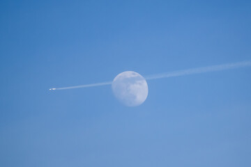 Full moon in the bright blue sky with a passing plane and a contrail in front. Plane passing by the moon