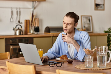 Young handsome man enjoys breakfast while working on laptop in a cozy kitchen setting