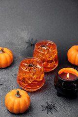 Two spooky amber drinks with ice and plastic spiders sit near mini pumpkins and cauldron candle