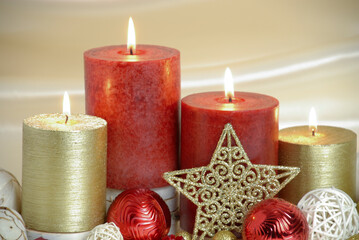Red and gold burning Christmas candles surrounded by natural potpourri elements and gold stars on a cream satin draped background