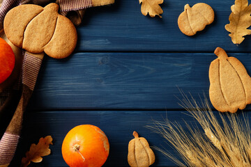 Autumn flat lay with pumpkin cookies, plaid cloth, pumpkins, wheat, and leaves on dark blue wood background.
