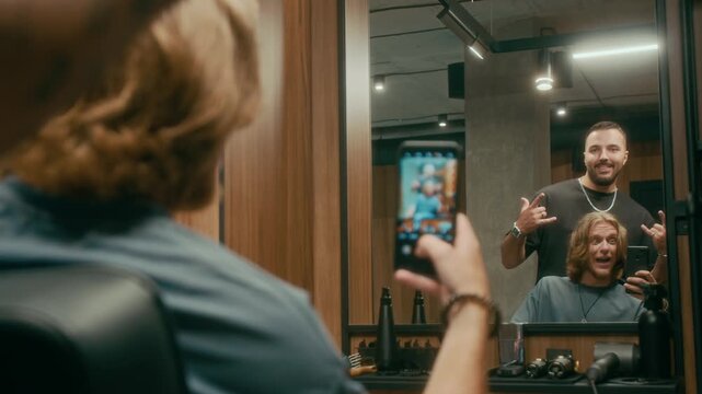 Medium rear shot of mirror reflection of cheerful young Caucasian guy with red hair taking selfies on smartphone with barber friend after getting new haircut, making funny faces and gestures