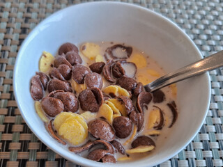 Bowl of mixed chocolate and corn cereal with milk, served with a spoon on a woven placemat, perfect breakfast or snack concept