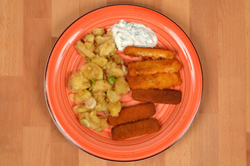A top-down shot of a meal with fish sticks, potato salad, and tartar sauce on a ribbed orange plate