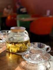 Transparent teapot with milk oolong leaves brewing, glass cup on the table. Blurred background with a couple at a cafe creates a cozy tea ceremony and lifestyle mood