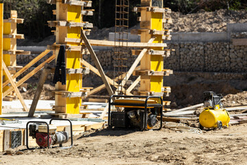 New building foundation work in progress. Shows yellow formwork, rebar, concrete columns, a water pump, portable generator, and an air compressor.