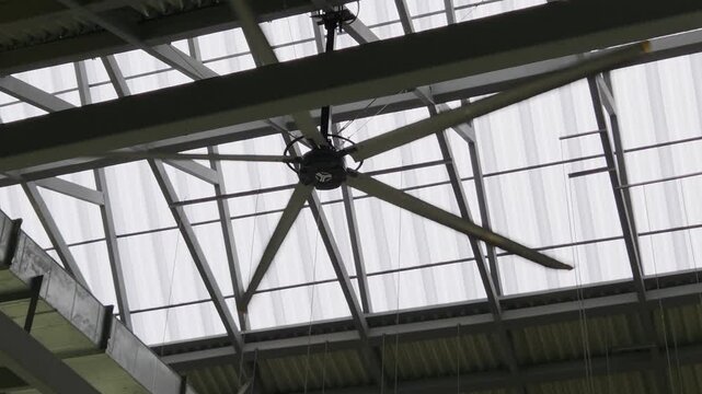 A low-angle shot of a large, industrial ceiling fan (HVLS fan) spinning and suspended beneath a sloped, translucent panel roof.