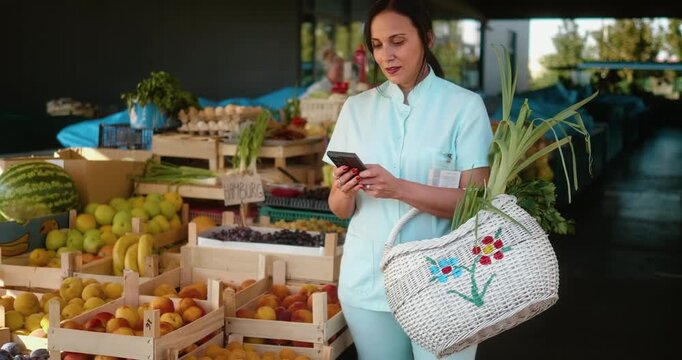 Caregiver or nurse in uniform at farmers market checking shopping list on mobile phone while buying fresh fruits and vegetables, placing produce into basket, concept of health and lifestyle. - Powered by Adobe