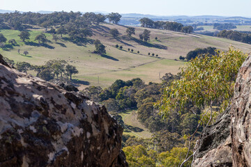 Scenic view from Hanging Rock in the Macedon Ranges, Victoria, Australia. Volcanic rocks in the foreground overlooking farmland, scattered trees, cows, distant hills, and the Hanging Rock Racecourse.