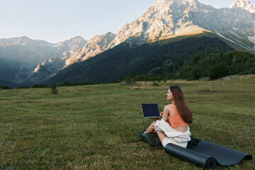 Fototapeta premium A woman sits on a rolled mat in a grassy meadow, using a laptop with a dramatic mountain backdrop. She smiles slightly, focused on work, creating a calm outdoor office mood.