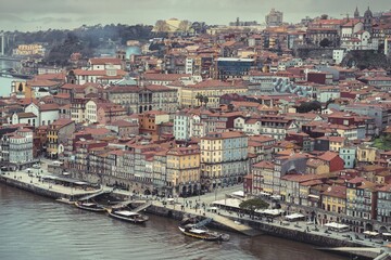 A cityscape shows historical buildings with red rooftops in Porto, Portugal. People walk along the riverfront, and boats are docked on the Douro River during the day