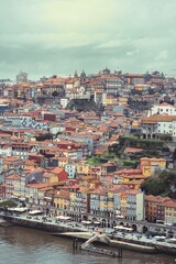 Obraz premium A wide view captures Porto, Portugal, with its cluster of buildings and red rooftops. A river flows at the edge of the town. The sky is covered with gray clouds on a late afternoon