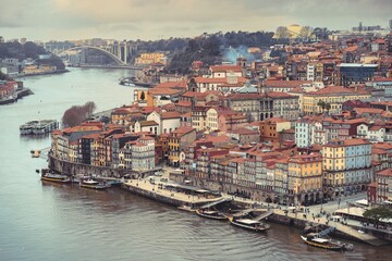 The Ribeira district in Porto presents its timeless charm. Buildings cluster along the Douro River, while boats dock and people stroll on a cloudy afternoon