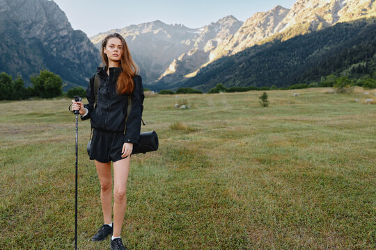A young woman stands in a mountain meadow with a trekking pole, wearing dark outdoor gear and a crossbody bag. She appears confident and ready for an adventurous outdoor day. - Powered by Adobe