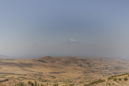 View of sun-baked hills stretch toward the distant, snow-capped peak under a hazy sky, creating a tableau of arid beauty, Kotayk Province, Armenia.