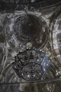 View of an ornate chandelier hanging beneath the aged, textured stone dome with intricate patterns and subtle lighting, Geghard Monastery, Mets Gilanlar, Ararat Province, Armenia.