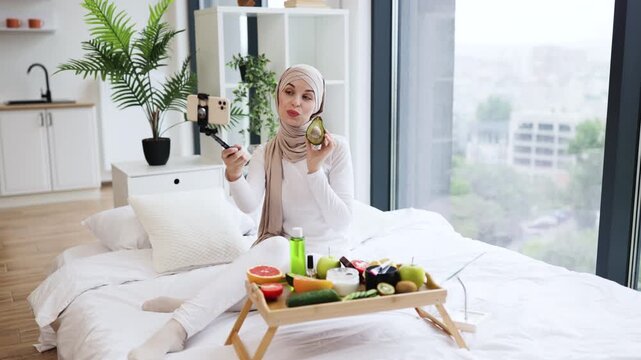 Young Muslim woman wearing hijab records skincare tips using avocado in bright modern room