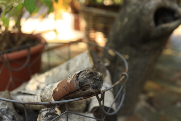 Wood logs are neatly arranged in a wire basket, surrounded by plants and a tree in a sunny garden setting. The scene captures a peaceful outdoor retreat.