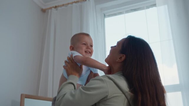 Medium shot shows mother joyfully lifting her two year old son in her arms in a bright room. A lively lifestyle moment full of movement, smiles, and everyday happiness at home. High quality 4k footage