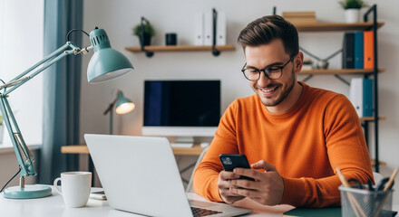 Smiling man in orange sweater happily using smartphone at desk with laptop and coffee, modern home office setup