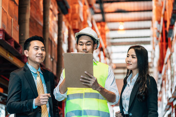 business man and woman in a suit smiles as he talks to a warehouse worker in a hard hat and safety vest, holding a clipboard. They are in a warehouse surrounded by tall shelves of merchandise
