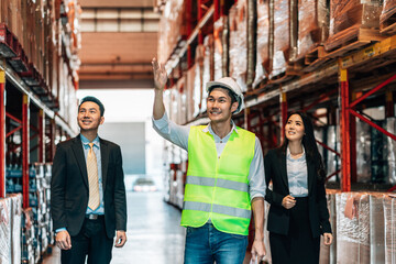 businessman and a businesswoman in suits walk through a warehouse with a warehouse worker in a hard hat and safety vest. The worker has his hand raised, gesturing toward the shelves as he explains