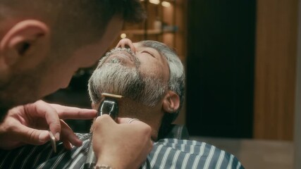 Close-up of face and hands of focused professional barber with tattoos styling beard for mature customer using electric trimmer and comb, in precise lines, during grooming appointment