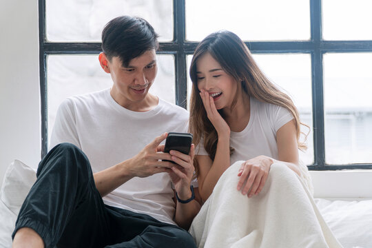 Cheerful asian couple using smartphone application browsing shopping online sitting together on comfortable couch at apartment. Husband wife smiling social network relax on couch. Lifestyle technology