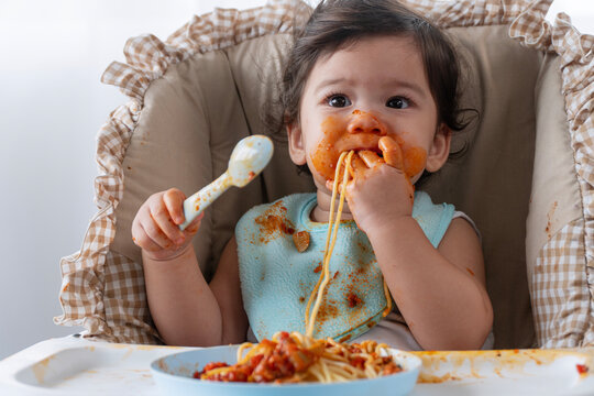 Adorable messy little child girl hungry use hand eating spaghetti sitting in high-powered chair at home. Toddler child with tomato sauce making mess face looking at food. Self-feeding concept