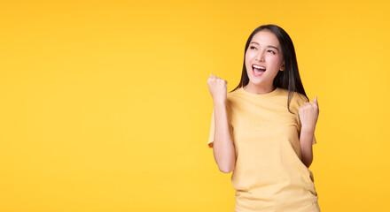 Joyful asian woman successful excitement up, keeps hands raised arm standing over isolated yellow background copy space. Excited young girl holding raised arm fist screaming succeed worker concept.
