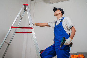 Professional male worker using a power drill while standing on a ladder in a modern interior space during home renovation project