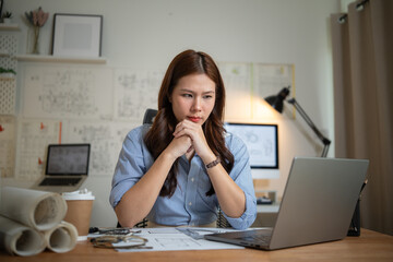 Focused female architect sitting at desk, looking thoughtful while analyzing project plans on laptop.