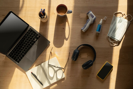 Remote work desk setup at home during pandemic. Laptop, smartphone, protective medical masks, hand sanitizer and electronic thermometer on table.