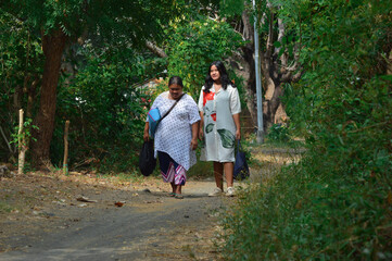 A woman and girl take a gentle walk along a shaded rural road, observing the greenery and enjoying calm conversation with supplies in hand.