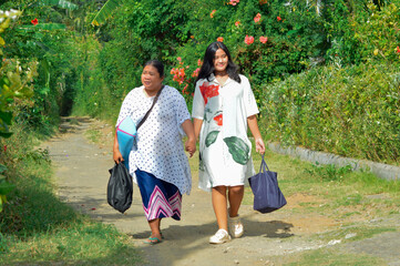 Fototapeta premium Two women enjoy their walk carrying supplies along a path bordered by blooming flowers, sharing a cheerful and peaceful rural moment together.