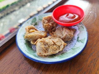 Tahu walik. Crispy browned tofu walik served in a natural wooden bowl with spicy sauce, with a blurry backdrop of a flowing river and rice fields. Menu in outdoor cafe.