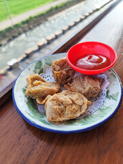 Tahu walik. Crispy browned tofu walik served in a natural wooden bowl with spicy sauce, with a blurry backdrop of a flowing river and rice fields. Menu in outdoor cafe.