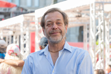 A man aged 50-55 is smiling in a blue shirt against the background of a city crowd.