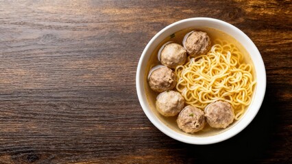 A bowl of flavorful beef meatballs in a light broth with noodles sits atop a dark wooden surface.