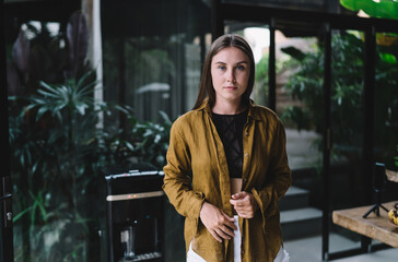 Young businesswoman standing calmly in front of glass wall, wearing mustard shirt over casual top, showing thoughtful and confident remote professional.