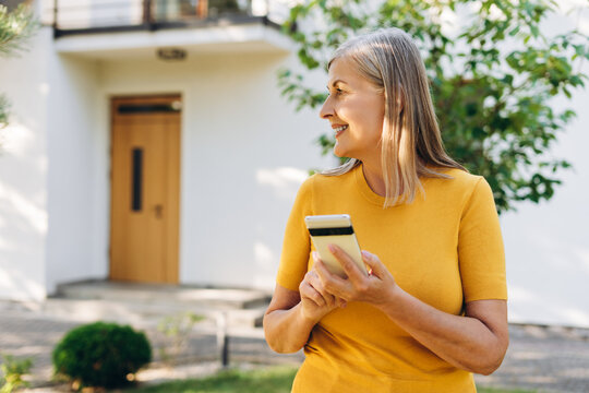 Smiling senior woman using smartphone text message, chatting in front of house - Powered by Adobe