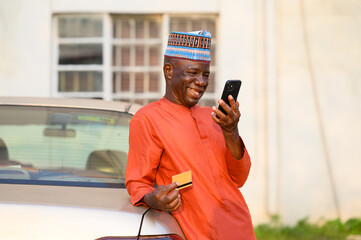 Elderly Nigerian man in traditional orange attire and cap smiling while holding a smartphone and a bank card, leaning against a car outdoors.