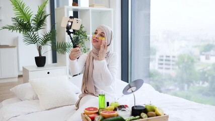 Young Muslim woman wearing hijab recording vlog about using gold under-eye patches. She sits in a bright bedroom with skincare products and fruits on display, sharing beauty tips with her audience. - Powered by Adobe