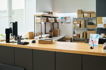 Empty modern mailroom counter displaying cardboard package on wooden surface with desktop computer and office supplies, shelves in background holding various parcels and shipping boxes