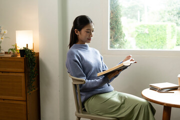 Young Asian woman enjoying a peaceful moment reading a book near window. Calm lifestyle, self-learning, and mindfulness at home.