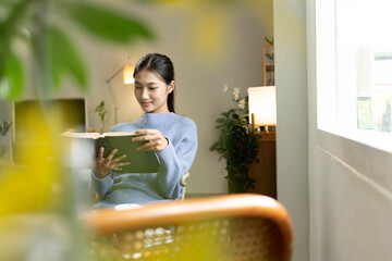Young Asian woman enjoying a peaceful moment reading a book near window. Calm lifestyle, self-learning, and mindfulness at home.