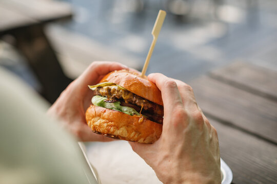 Close-up of a man sitting at a garden table eating a juicy cheeseburger with pickles and salad