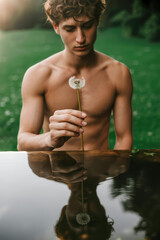 Contemplative Young Man Holding a Dandelion Over Still Water with Lush Green Background
